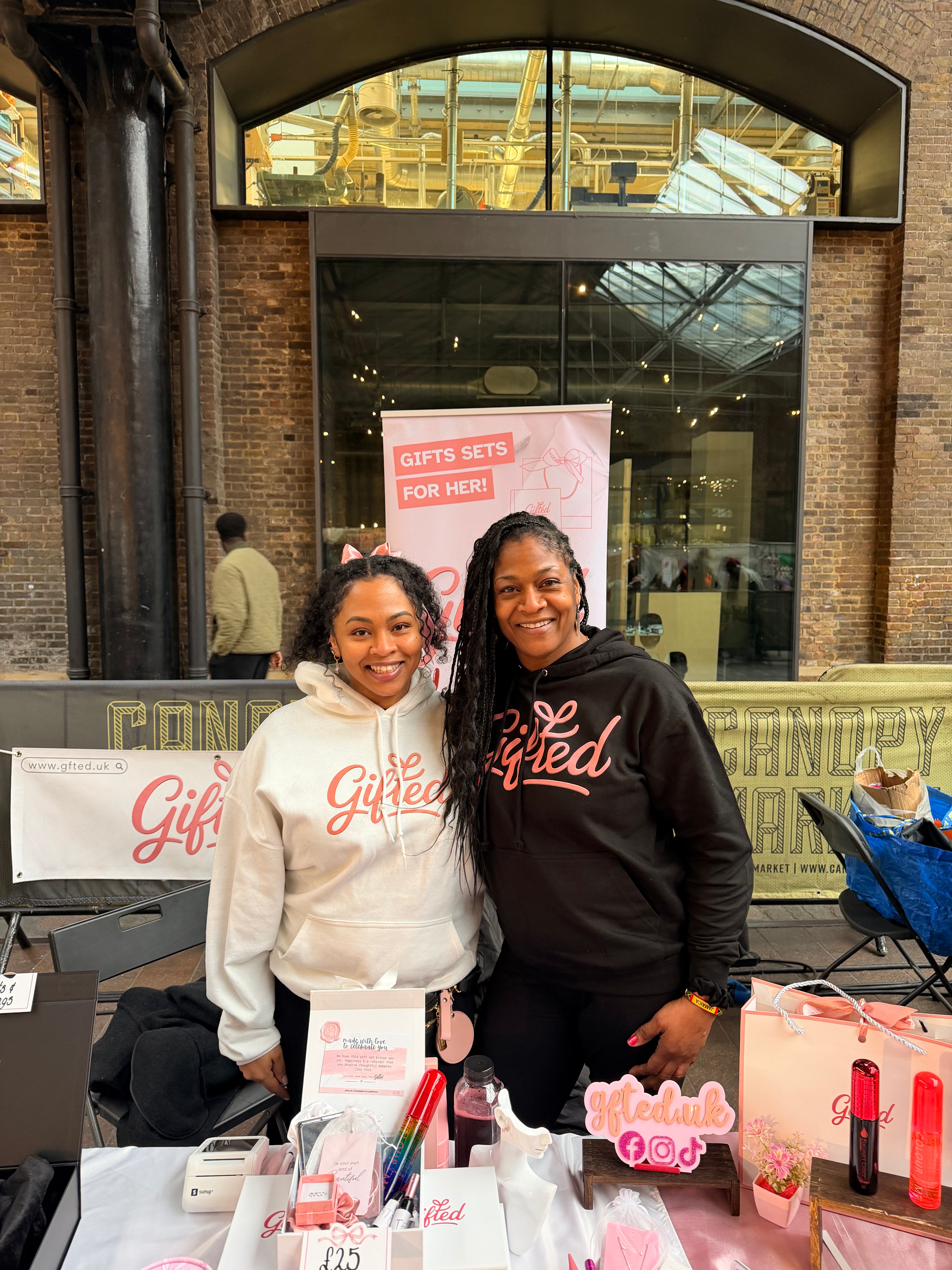 Two women standing behind a Gifted market stall table with promotional materials in an indoor setting.