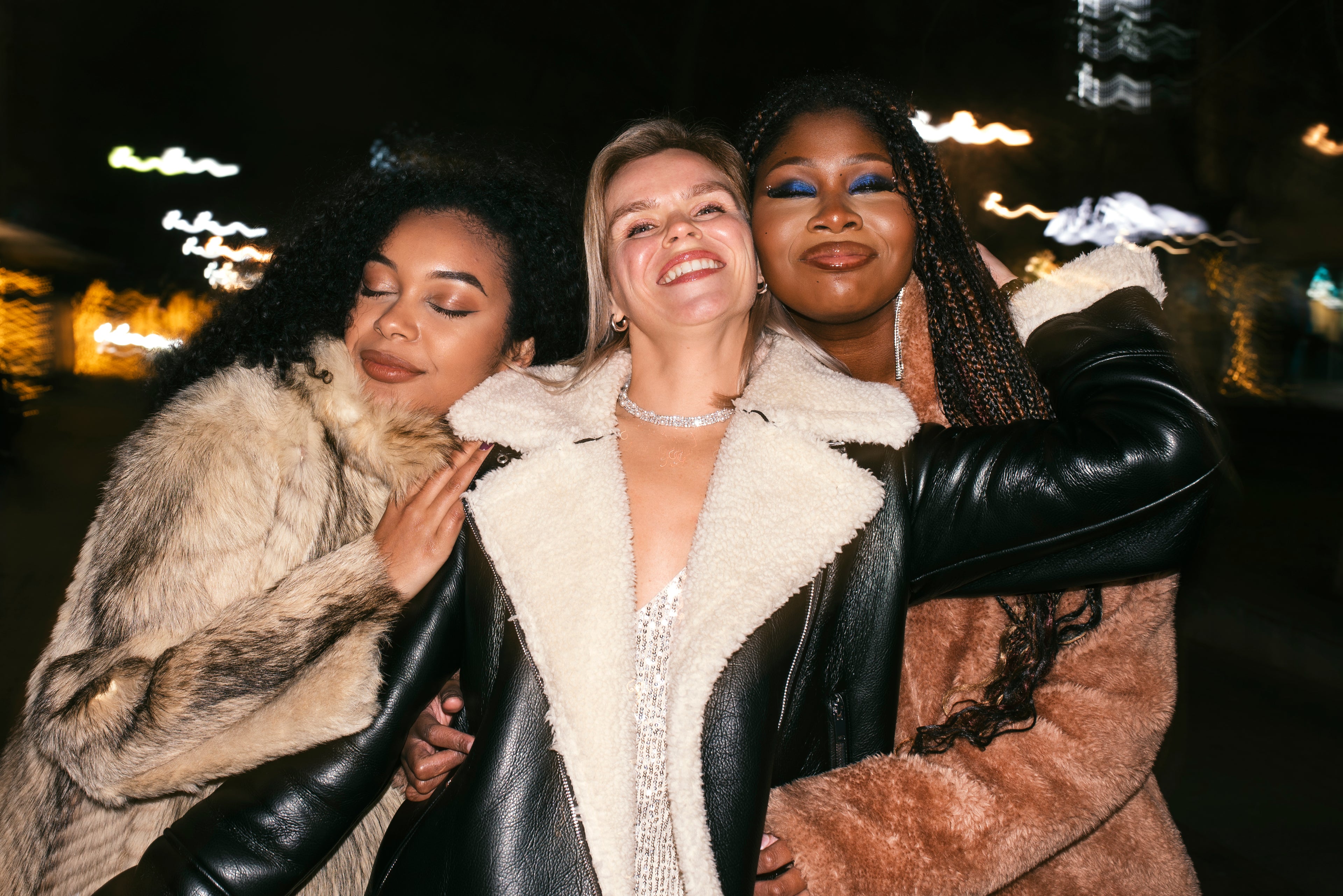 Three women posing together outdoors at night, wearing stylish winter coats.