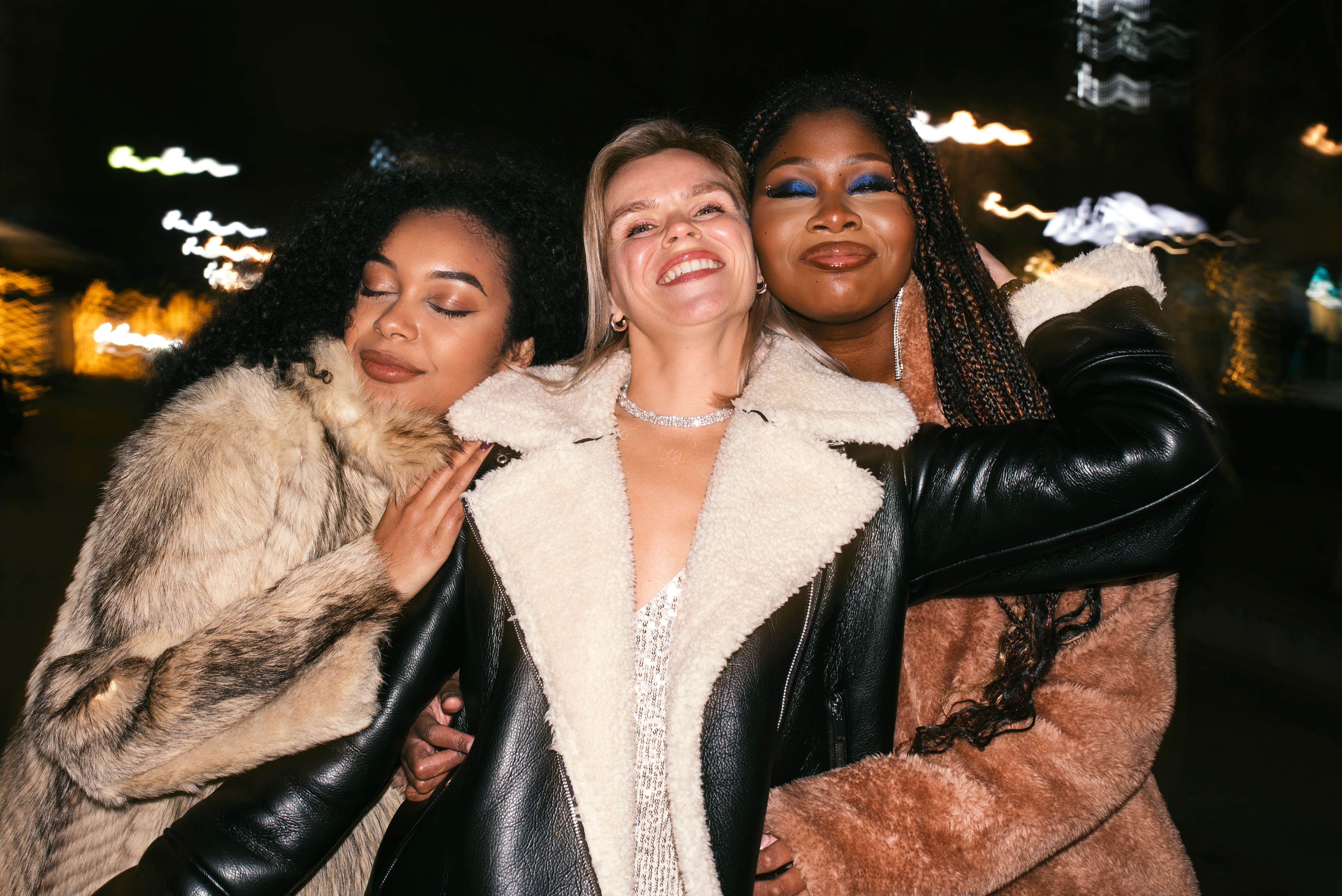 Three women posing together outdoors at night, wearing stylish winter coats.