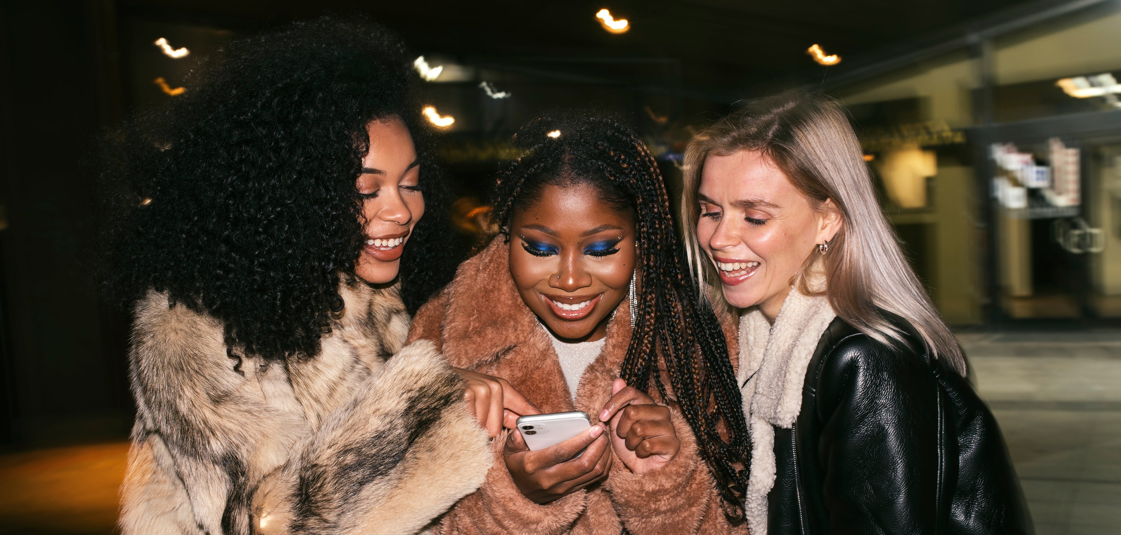 Three women in winter coats looking at a phone together in a dimly lit outdoor setting.